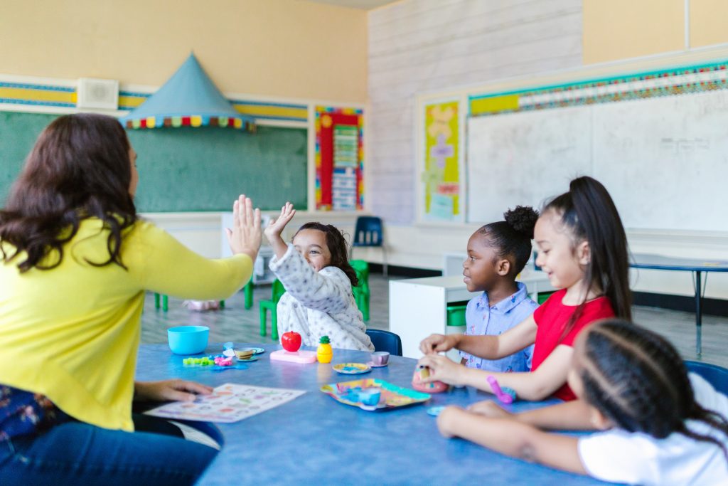 Eine Lehrerin sitzt 4 Mädchen im Grundschulalter gegenüber. Einem lachenden Mädchen gibt sie High Five.